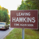 A car on a road passes by a red sign with white text that reads "Leaving Hawkins Come Again Soon"